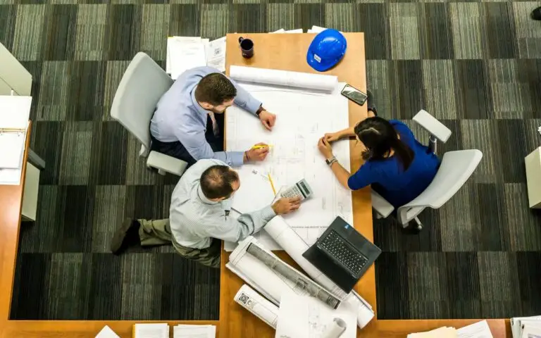 Three engineers collaborating over architectural blueprints on a table in an office, with a hard hat, laptop, and calculator nearby