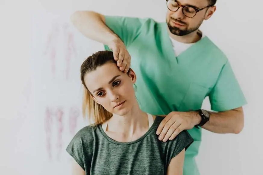 A chiropractor in green scrubs examines a patient's neck and shoulder in a clinical setting