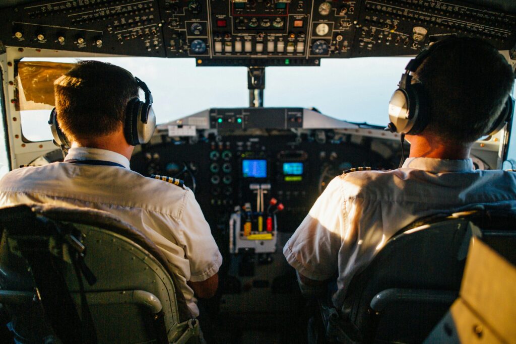 Two pilots in a cockpit operating an aircraft, viewed from behind, with a detailed control panel and screens in front of them