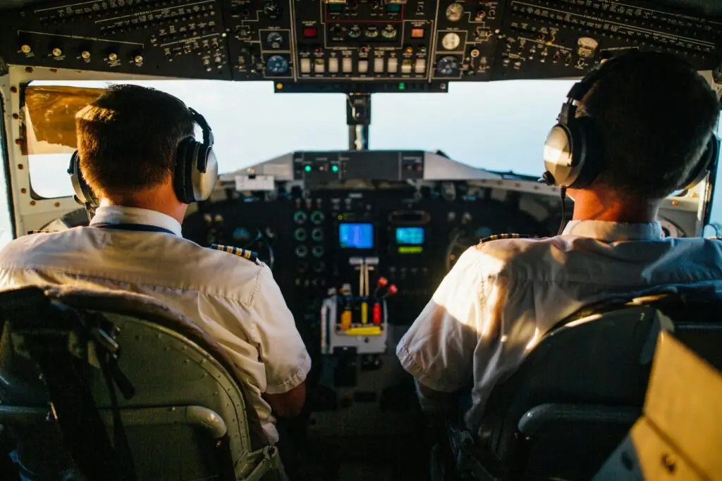 Two pilots in a cockpit operating an aircraft, viewed from behind, with a detailed control panel and screens in front of them