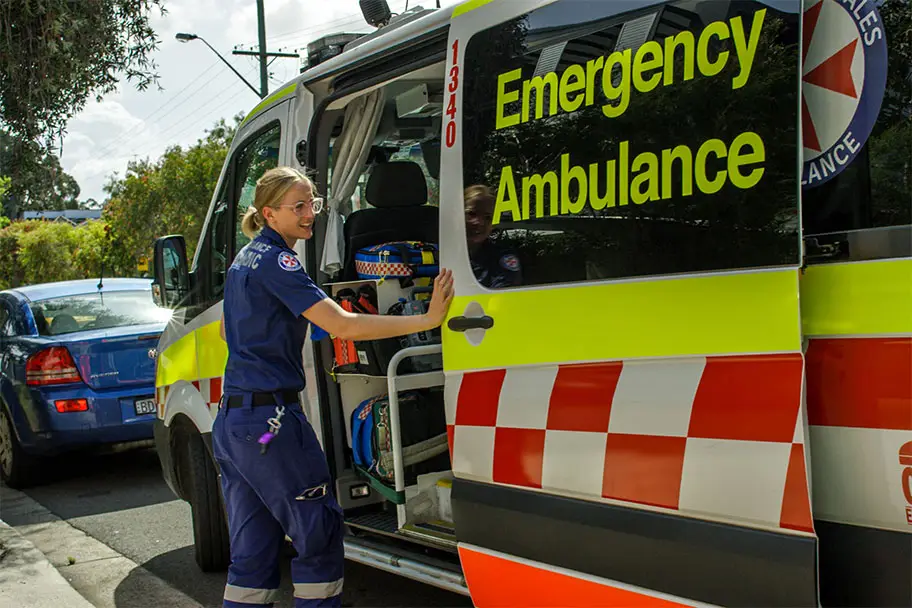 An ambulance officer in uniform stands next to an ambulance, preparing for a call